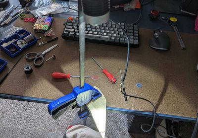 A cluttered desk workshop with a Dremel clamped upright over a keyboard and mouse, surrounded by polishing bits, screwdrivers, and wire